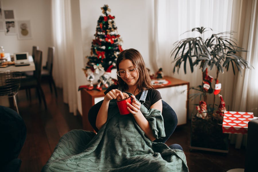 Devojka sedi u fotelji pokrivena ćebetom i umače keks u crvenu &scaron;olju