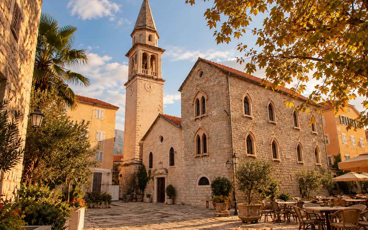 Pogled na staru kamenu crkvu s tornjem na sunčanom trgu okruženom kafićima.